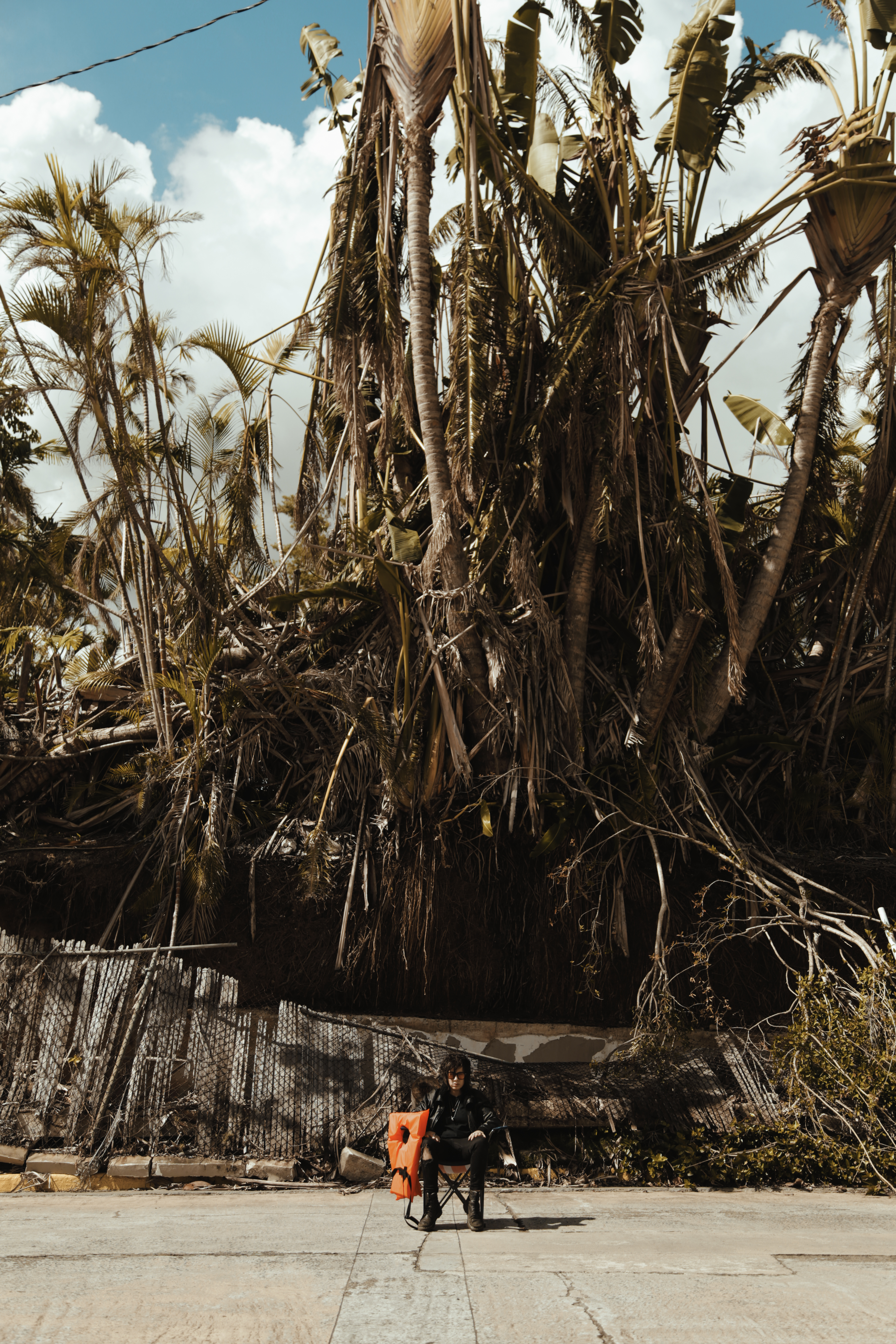 In 'Welcome to Paradise I,' Jo Cosme sits in front of destroyed fence and trees while staring into the camera next to a life jacket