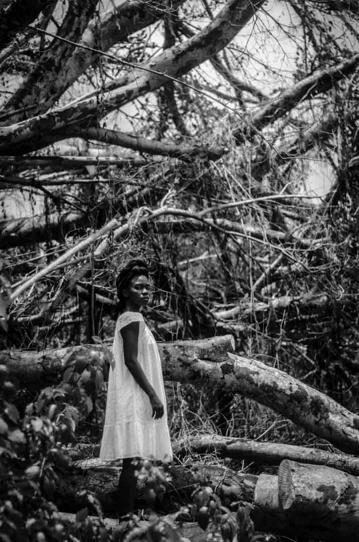 A young Black woman stares over her shoulder into the camera while she stands among trees destroyed by the storm in David Berg's black-and-white photograph, Regrowth I